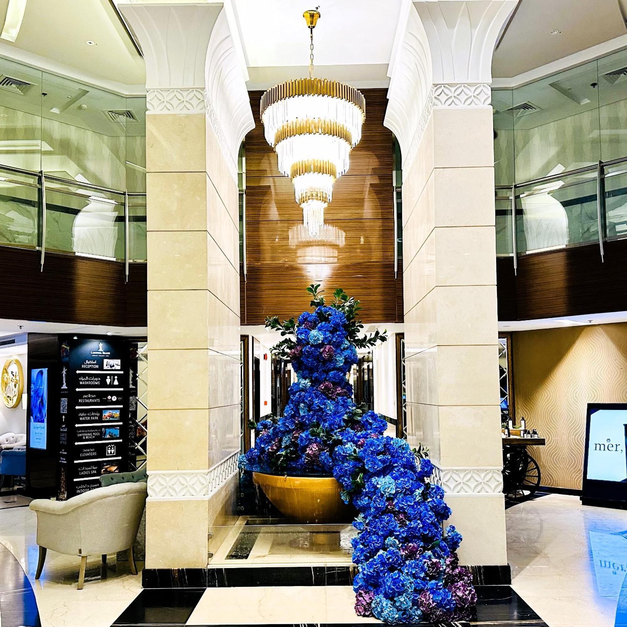 Lagoona Beach Luxury Resort And Spa Manama Exterior foto The photo depicts an elegant lobby or entrance area of a building. Central to the scene is a large floral arrangement featuring vibrant blue flowers. This arrangement is positioned in a decorative bowl. Above it hangs a stylish chandelier with a laye