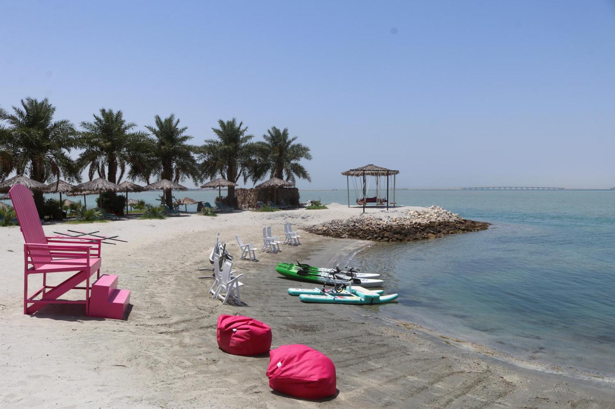 Lagoona Beach Luxury Resort And Spa Manama Exterior foto The photo depicts a serene beach scene with clear blue water and a sunny sky. In the foreground, there are colorful pink bean bags and a large pink chair on the sand. A few white chairs and loungers are positioned near the water's edge. To the side, 