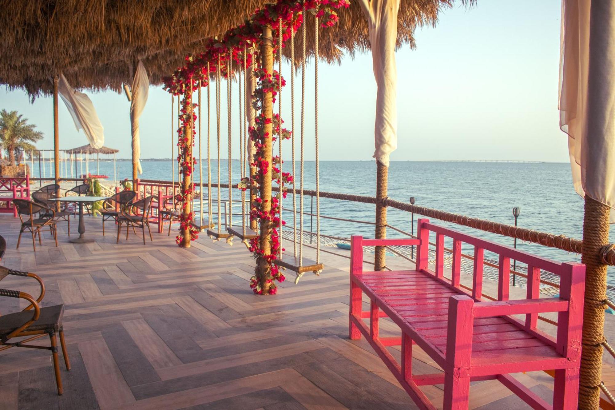 Lagoona Beach Luxury Resort And Spa Manama Exterior foto The photo depicts a scenic outdoor area by the water, with a wooden deck and a thatched roof. Two swings adorned with flowers are hanging from the ceiling, providing a charming touch to the space. In the foreground, there is a bright pink bench, addi