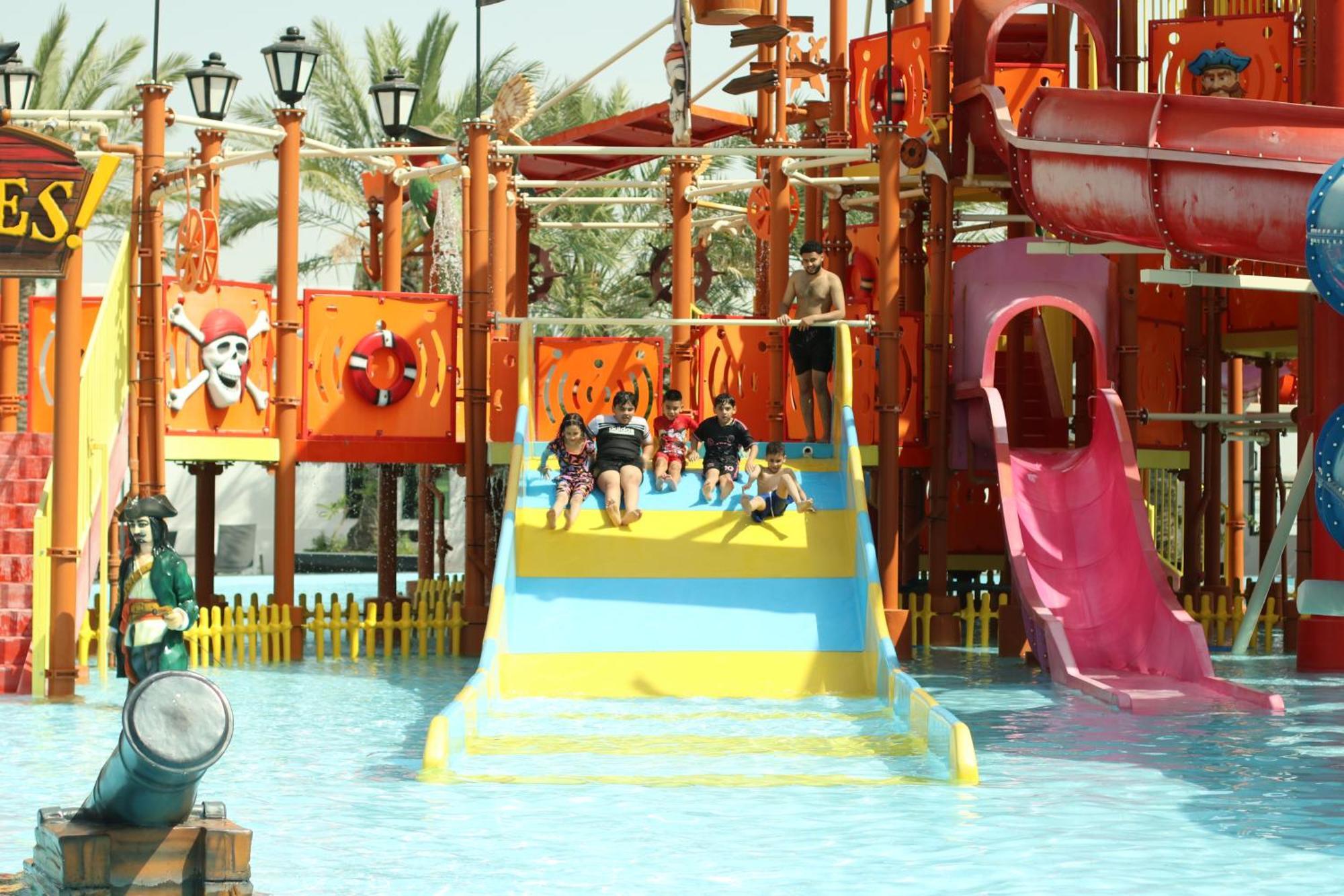 Lagoona Beach Luxury Resort And Spa Manama Exterior foto The photo shows a colorful water park scene featuring a play area with multiple slides. There are children enjoying themselves, sliding down bright yellow and pink slides into a pool of water. The structure has pirate-themed decorations, including a 