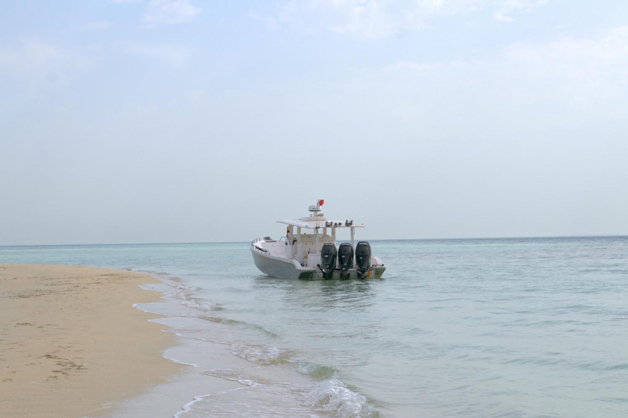 Lagoona Beach Luxury Resort And Spa Manama Exterior foto The photo depicts a small boat anchored near a sandy beach, partially submerged in calm, clear water. The boat is white and features three outboard motors on its stern. In the background, the sea stretches out toward the horizon under a light, cloudy