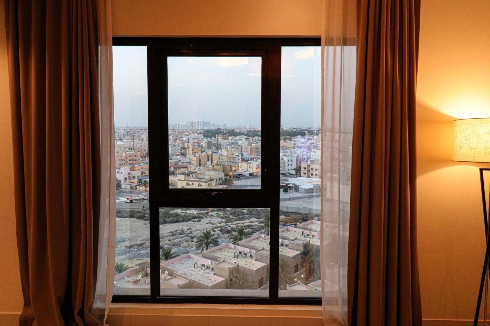 Lagoona Beach Luxury Resort And Spa Manama Exterior foto The photo shows a window with a black frame and sheer curtains. Through the window, there is a view of a cityscape, displaying a densely populated area with various buildings and rooftops. The sky appears slightly overcast, suggesting a muted lightin