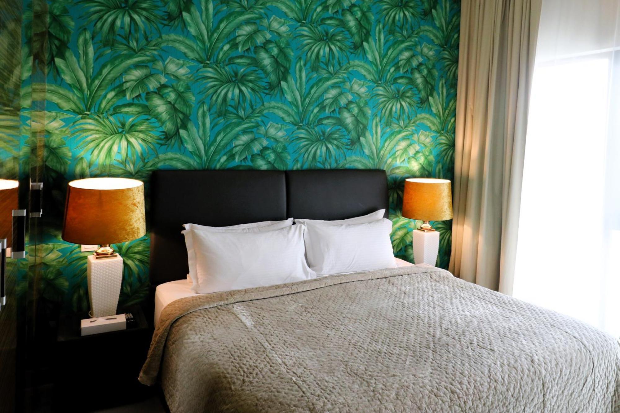 Lagoona Beach Luxury Resort And Spa Manama Exterior foto The photo shows a stylish bedroom featuring a large bed with white pillows and a gray blanket. The headboard is upholstered in black, providing a modern touch. Behind the bed, there is a vibrant green wallpaper with a tropical leaf pattern, adding a 